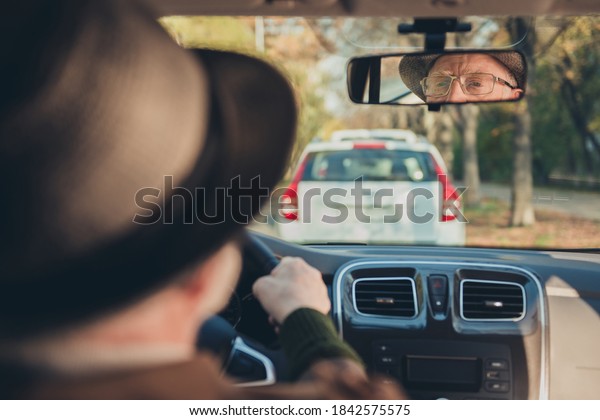 Photo of retired old man drive sit seat reflecting\
rare view mirror look road wear brown jacket headwear spectacles\
inside car