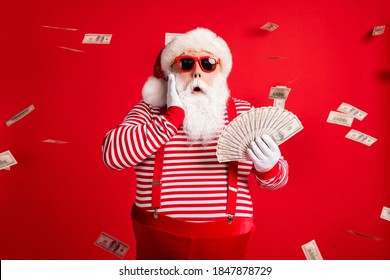 Photo of retired old man grey beard hold cash fan hand cheek shock money wear santa x-mas costume suspenders sunglass gloves striped shirt cap isolated red color background