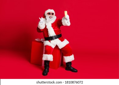 Photo of retired old man white beard sit giftbox show v-sign make selfie portrait stick-out tongue wear x-mas santa costume glove coat belt sunglass cap boot isolated red color background - Powered by Shutterstock