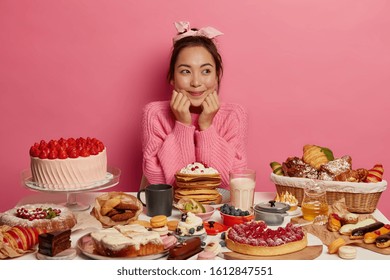 Photo Of Satisfied Asian Woman Sits At Table Overloaded With Many Desserts, Breaks Diet, Has Unhealthy Nutrition, Eats Homemade Cakes, Croissants, Dreams About Eating Sweets And Not Putting Weight