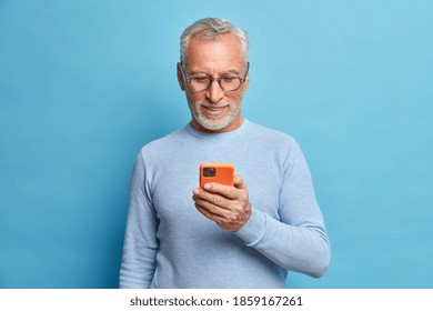 Photo of satisfied bearded man focused in smart phone surfs internet sends text messages in social networks uses modern technologies wears casual blue jumper poses indoor. Senior freelancer.