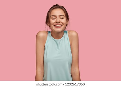 Photo Of Satisfied European Young Woman With Pleasant Broad Smile, Keeps Eyes Closed, Dreams About Something Pleasant, Shows Bare Shoulders, Dressed In Casual Vest, Isolated Over Pink Background