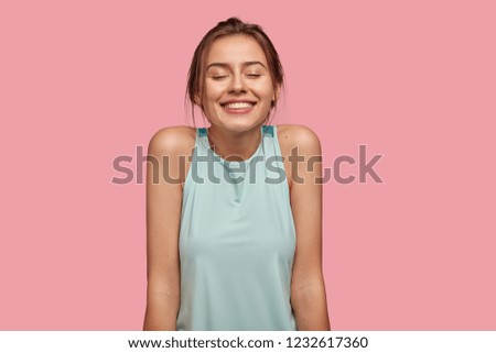 Photo of satisfied European young woman with pleasant broad smile, keeps eyes closed, dreams about something pleasant, shows bare shoulders, dressed in casual vest, isolated over pink background