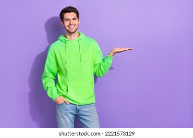 Photo Of Satisfied Guy With Brunet Hairdo Dressed Green Hoodie Hand Pocket Demonstrating Empty Space Isolated On Purple Color Background