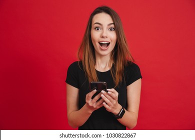 Photo Of Satisfied Woman In Black Clothing Looking On Camera While Using Cell Phone With Joy Over Red Background