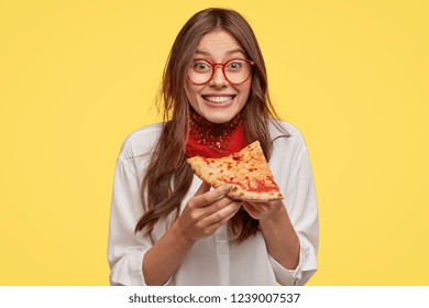 Photo of satisfied woman holds piece of pizza, feels pleased as spends free time with friends in pizzeria, looks happily directly at camera, wears casual outfit, isolated over yellow wall. Lunch