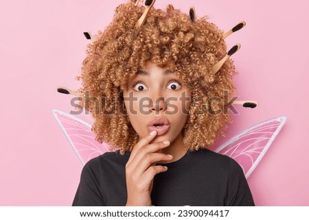 Photo of scared curly haired female tooth fairy with toothbrushes in hair keeps mouth opened and stares speechless finds out something horrible comes on costume party poses against pink background