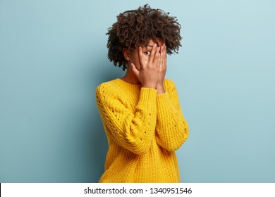 Photo of scared ethnic woman peeks through fingers, hides face, afraids of seeing something terrible, has Afro haircut, wears yellow clothes, isolated over blue wall. Omg, I dont want to see it