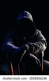 Photo Of Scary Horror Stranger Stalker Man In Black Hood And Clothing Sitting On Chair On Dark Background.