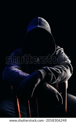 Photo of scary horror stranger stalker man in black hood and clothing sitting on chair on dark background.