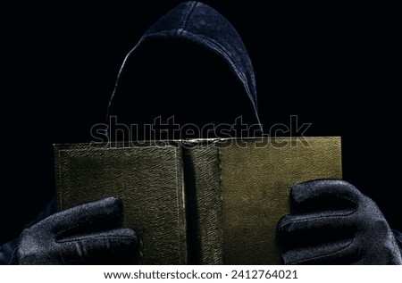 Photo of scary shaded hooded man holding opened book and peaking on dark background. Horror book concept.