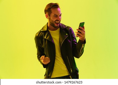 Photo Of Screaming Young Stylish Man Isolated Over Light Green Background Wall With Led Neon Lights Dressed In Leather Jacket Using Mobile Phone Make Winner Gesture.