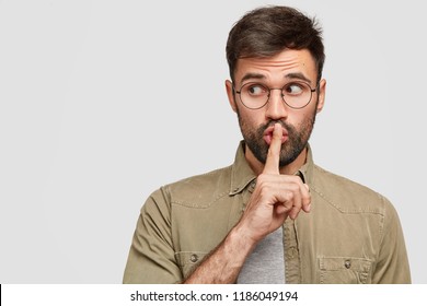 Photo Of Secret Young Man Makes Shush Gesture, Looks Surprisingly Aside, Notices Something Strange, Has Dark Stubble, Isolated Over White Background With Copy Space For Your Promotional Text