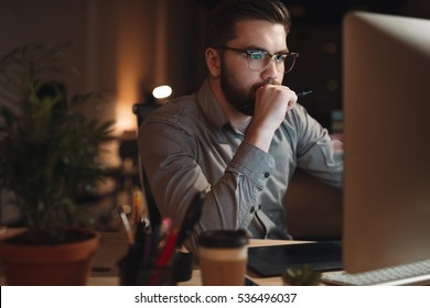 Photo Of Serious Bearded Web Designer Dressed In Shirt Working Late At Night And Looking At Computer. Holding Pen.