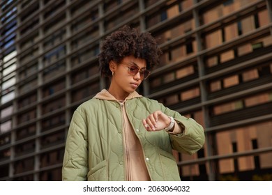 Photo Of Serious Curly Woman Hurries For Work Or Meeting Looks Attentively At Wrist Watch Checks Time Wears Trendy Sunglasses And Jacket Poses Outdoors Tracks Clock. People And Lifestyle Concept