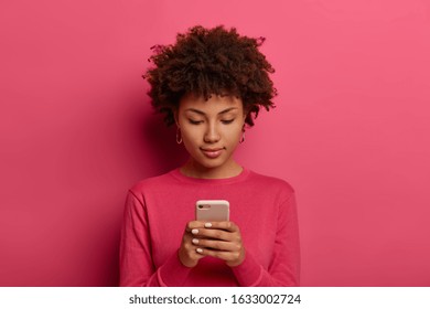 Photo of serious curly woman uses modern device, orders taxi via smartphone application, texts or browses in social media, wears bright crimson clothes, isolated on pink background, checks email box - Shutterstock ID 1633002724