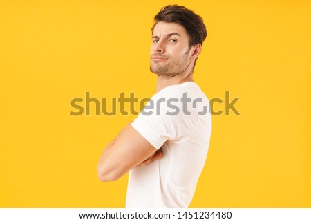 Photo of serious man in basic t-shirt frowning and looking at camera with arms crossed isolated over yellow background