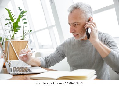 Photo of serious mature man making notes while working with laptop and cellphone in cafe indoors