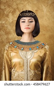 Photo Of Serious Woman With Cleopatra Makeup, Closeup Portrait Of Beautiful Female With Stylish Haircut Agaist Golden Background, Young Lady Wearing Fashionable Golden Necklace, Beauty Salon