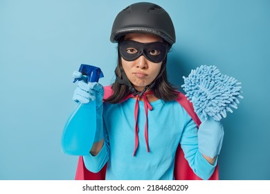 Photo of serious woman wears black protective helmet eyemask and rubber gloves dressed in superhero costume holds spray detergent and rag going to wash everything dirty in room isolated over blue
