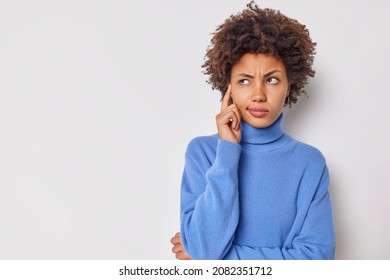 Photo of serious young curly woman keeps finger on temple tries to concentrate considers something contemplates about important thing wears casual blue jumper isolated over white background. - Shutterstock ID 2082351712