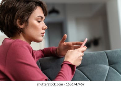 Photo of serious young woman in leisure clothes using smartphone while sitting on sofa in living room - Shutterstock ID 1656747469