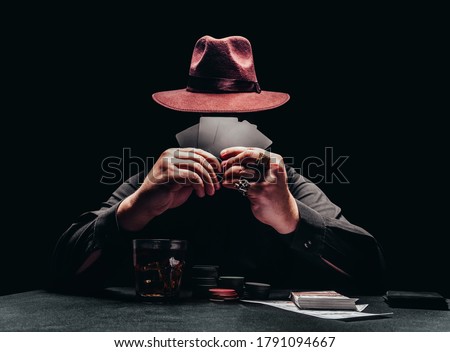 Photo of a shaded gangster in black shirt and hat playing poker and holding black cards with game chips and money.