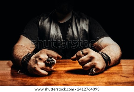 Photo of a shaded male muscular biker in leather jacket and rings sitting by a wooden table.