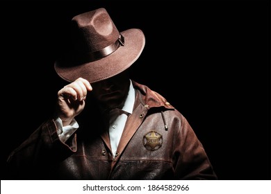Photo of a shaded sheriff officer with badge in jacket putting on cowboy hat on black background.