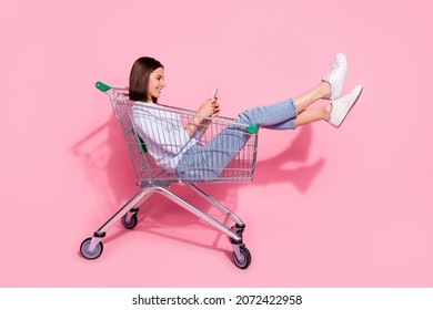 Photo of shiny cute young woman wear white sweater sitting shopping cart chatting modern gadget smiling isolated pink color background