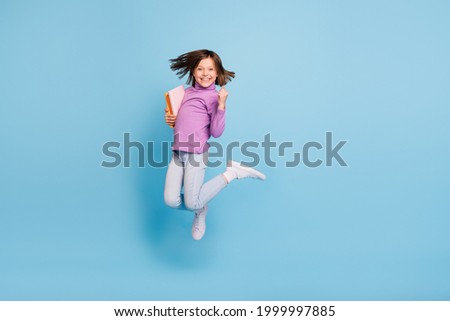 Photo of shiny lucky school girl wear purple turtleneck jumping holding copybooks rising fist smiling isolated blue color background