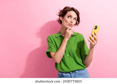 Photo of shiny unsure young lady wear green shirt typing apple iphone samsung device arm chin empty space isolated pink color background