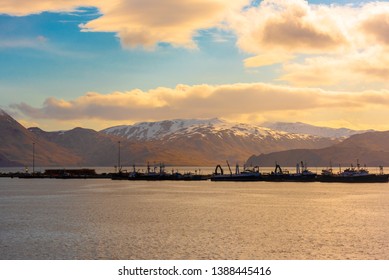 Photo of ships silhouetted against snow-capped mountains, blue sky and gold tinged clouds in Dutch Harbor, Alaska.