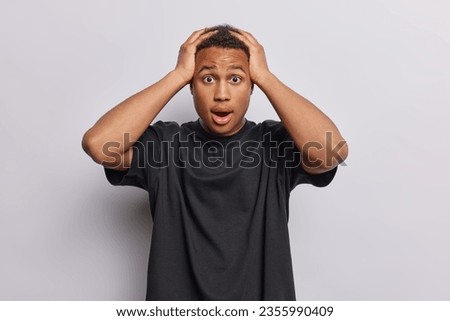 Photo of shocked dark skinned young man keeps hands on head feels embarrassed reacts to something terrible wears casual black t shirt isolated over white background. People and reactions concept