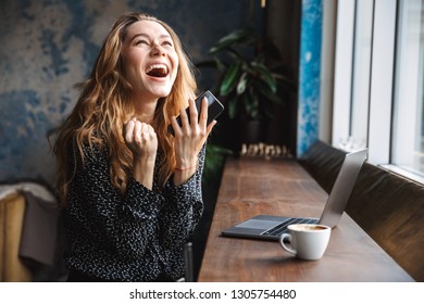 Photo Of Shocked Excited Beautiful Young Pretty Woman Sitting In Cafe Indoors Using Laptop Computer And Mobile Phone.