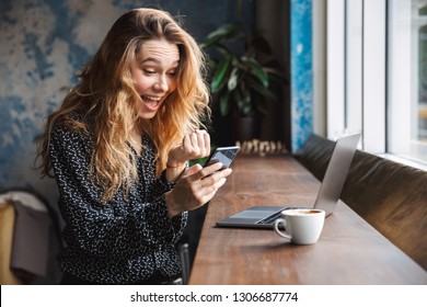 Photo Of Shocked Excited Beautiful Young Pretty Woman Sitting In Cafe Indoors Using Laptop Computer And Mobile Phone.