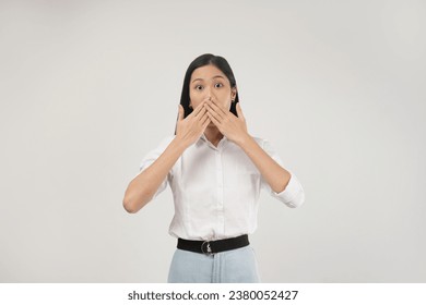 Photo of shocked young asian woman shuts her mouth with startled, impressed face expression, stands over white background.
