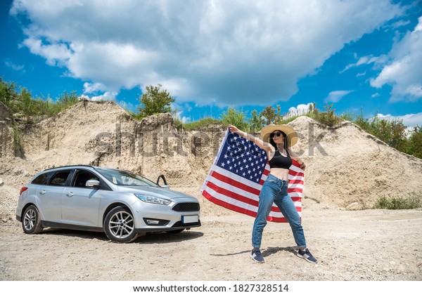 Photo shoot of a charming lady with the\
USA flag near the car in a sand quarry in\
summer