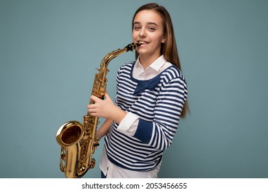 Photo shot of beautiful happy smiling brunette little girl wearing striped longsleeve standing isolated over blue background wall playing saxophone looking at camera