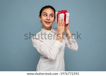 Photo shot of beautiful happy smiling brunette young woman isolated over blue background wall wearing white shirt holding white gift box with red ribbon and looking to the side