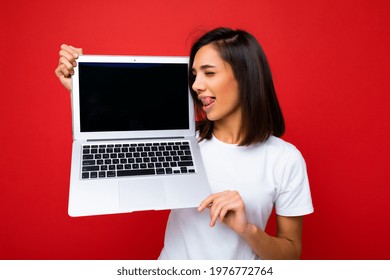 Photo Shot Of Funny Smiling Beautiful Happy Overjoyed Young Woman With Short Brunet Hair Crazy Sexy Tongue And Close Eyes Holding Computer Laptop Wearing White T-shirt And Jeans Isolated Over Red Wall