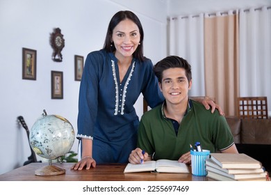 Photo shot of Indian teenager son and young mother both are smiling together. Indian teenager son and young mother both are smiling while doing homework at home, mummy standing near her seated son