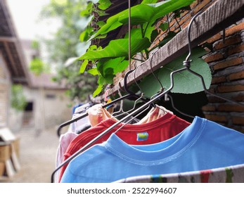 The photo shows a close-up of clothes hanging on a clothesline outside. The clothes are a blue shirt, a red shirt, and a brown shirt. The clothesline is in front of a brick wall and some green leaves. - Powered by Shutterstock