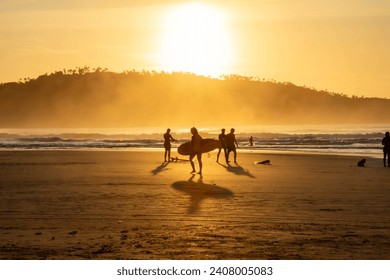 The photo shows the flat, sandy beach of Campeche on Santa Catarina Island at dawn. People and some Surfers are walking along the shore. In the background, you see the sun rise above Campeche Island. - Powered by Shutterstock