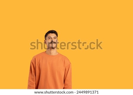 Photo shows a millennial guy with a moustache, standing against a vivid orange isolated background, giving a funny expression, copy space
