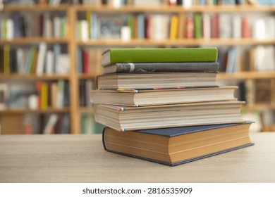 The photo shows a neat stack of five hardcover books resting on a light wooden table in what appears to be a quiet library or bookstore. The books vary slightly in thickness, with mostly neutral covers in beige, white, blue, and brown tones. Their pages are clean, creating a tidy and well-kept appearance. Behind the stack, large bookshelves filled with many colorful books stretch across the background. The shelves and books are intentionally blurred, producing a shallow depth of field that draws attention to the foreground. Warm, natural-looking lighting gives the scene a welcoming, calm atmosphere, suggesting a comfortable place for reading, studying, or research. 
