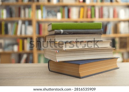 The photo shows a neat stack of five hardcover books resting on a light wooden table in what appears to be a quiet library or bookstore. The books vary slightly in thickness, with mostly neutral covers in beige, white, blue, and brown tones. Their pages are clean, creating a tidy and well-kept appearance. Behind the stack, large bookshelves filled with many colorful books stretch across the background. The shelves and books are intentionally blurred, producing a shallow depth of field that draws attention to the foreground. Warm, natural-looking lighting gives the scene a welcoming, calm atmosphere, suggesting a comfortable place for reading, studying, or research. 
