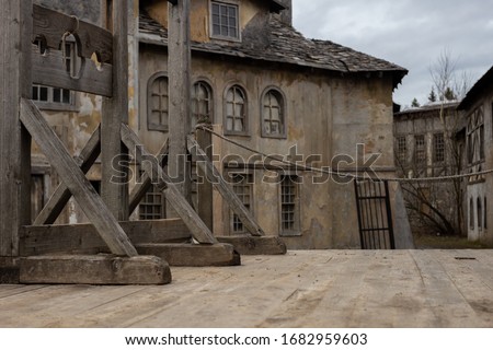 The photo shows part of a dark medieval or pirate town with a guillotine on a wooden platform. In the background, houses with broken windows, desolation and destruction as during the plague pandemic.