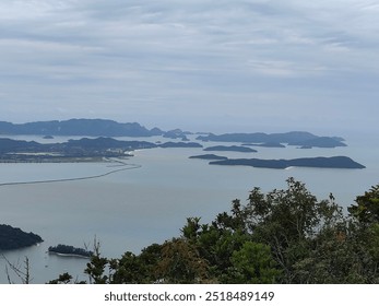 The photo shows a scenic view of the sea dotted with small islands under a cloudy sky. Lush greenery in the foreground contrasts with the calm water and distant mountains on the horizon. - Powered by Shutterstock