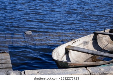 The photo shows a small boat docked at a pier by the water. The water has a rich blue hue, and a portion of the wooden pier is almost fully submerged, creating beautiful reflections on the surface. Th - Powered by Shutterstock